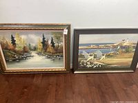 Two framed paintings sitting on floor against wall, one river scene and one coastal scene.