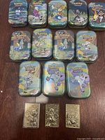 Front view of 15 collectible Pokemon tins with various character artwork, plus 3 gold plated Pokemon cards laid out below.
