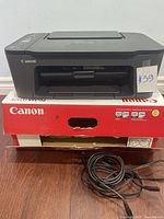 Front view of Canon PIXMA printer showing front paper output tray and Canon branding. Printer sits on red Canon box with ink cartridge model numbers visible on the box.