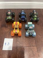 Top down photo showing all 5 small monster trucks arranged on a wooden floor with numbered tag #3 visible.