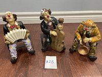 Three vintage clown figurines playing musical instruments placed on the floor with a #28 paper in front of them.