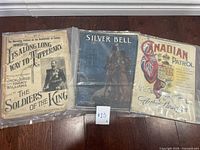 Three vintage music sheets displayed side by side on a wooden floor. Titles visible: 'It's Along Long Way to Tipperary,' 'Silver Bell,' and 'Canadian Patrol.'