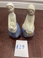 Two vintage porcelain figurines of ladies with scarves and blue skirts, placed on a wood floor, alongside a card marked '#29'.