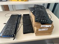 Three keyboards displayed on books and cardboard boxes including 2 wired black keyboards and 1 slim keyboard.