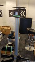 Full view of the standing TV/computer stand showing top mount with arrow decals, vertical silver support column and black base with caster wheels.