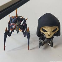 Two metal and plastic figurines on white surface - a Reaper with hood and dual guns, and a purple-orange colored Zerg unit with multiple legs and spikes