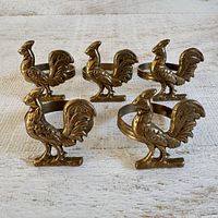 Front angled view of five brass rooster napkin rings showcasing their detailing and ring bands.