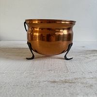 Front side view of the copper planter pot showing rounded body, black metal curved feet, and twisted handle.
