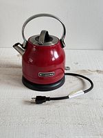 Red KitchenAid electric kettle side view showing handle, spout, and attached power cord with plug.