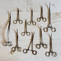 Top-down photo showing 9 metal surgical tools including hemostats and forceps laid on textured surface with visible signs of use, some curved and some straight tips.