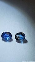 Pair of oval blue sapphires placed on a light surface with shadow indicating lighting conditions, showing color and clarity details.