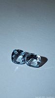 Two cushion cut blue topaz gemstones with clear facets and sky blue color lying side by side on a white surface.