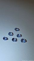 Six pear shaped light denim blue sapphires arranged on white background showing facets and natural color under lighting.