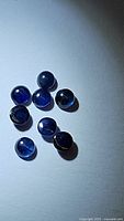 Eight natural blue sapphire round cabochon gemstones displayed on a white surface, showing their deep blue color and rounded smooth shape.