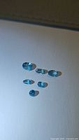 Six lagoon blue natural Cambodian zircon gemstones displayed on white surface, showing size and colors.