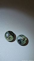 Two natural moss agate round cabochons on a textured grey surface, showing translucent greenish bodies with black moss-like inclusions.