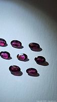 Eight natural oval cut rhodolite garnet gemstones in deep fuchsia to purple-pink color arranged on a white surface, clearly showing oval cuts and polished facets.