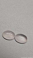 Two oval cabochon rose quartz gemstones placed on a grey textured surface, showing translucent pink color and smooth polished domed surface.