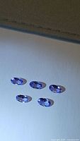 Five oval cut purple-blue tanzanite gemstones laid out on white paper, showing faceted tops and clarity.