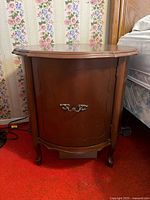 Front view of one wooden bedside table with curved legs, scalloped front panel, and metal drawer pull, placed beside a bed with floral wallpaper behind it.