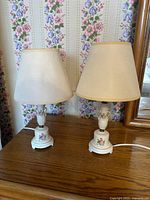 Two white ceramic table lamps on wooden surface against floral wallpaper, each with beige fabric shade and floral painted bases