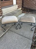 Photo showing two beige cushioned foot stools with black rod iron curved legs placed outside on concrete patio near steps and brick wall.