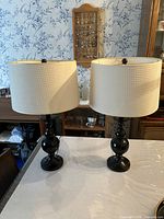 Pair of matching black table lamps with white textured fabric drum shades on a wooden surface with a background of wooden furniture and wallpaper.