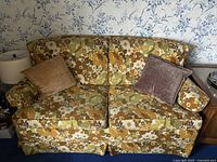 Front view of one floral patterned retro love seat with two square pillows in beige and brown velvet-like fabric and floral round pillow.