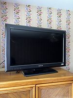 Front view of the black framed Sony Bravia TV on a wooden cabinet with floral wallpaper background, screen off.