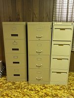Photo of three tall beige metal filing cabinets with four drawers each, placed side by side against a wood-paneled wall with a curtain.