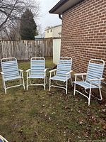 Four light blue plastic and white metal frame folding outdoor chairs arranged on grass near a brick wall and wooden fence.