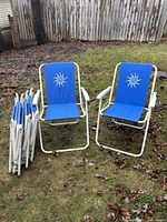 Six blue folding outdoor fabric chairs, two set up and four folded, shown outdoors on grass with rustic wooden fence in background.