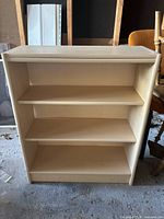 Front view of small bookshelf with three shelves, placed on concrete floor with various other furniture items around it.