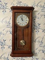 Full view of Bulova wooden wall clock with glass front and brass pendulum on floral wallpaper background.