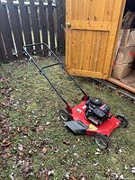 Red Canadiana push lawn mower outside near wooden shed, showing general view and handle