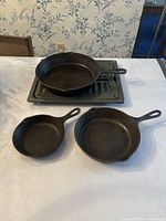 Photo shows three cast iron pans and one rectangular cast iron oven tray placed on a white surface against a wallpapered background.