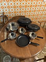 Photo showing four stainless steel cooking pots with glass lids, one black non-stick frying pan, and two large covered roasters on a wooden table