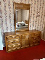 Front view of wooden dresser with six drawers and large matching mirror above it, placed against floral wallpaper covering the wall.