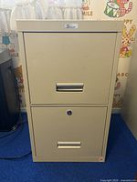 Front view of beige 2 drawer metal filing cabinet with silver handles and lock on lower drawer, placed on blue carpet with colorful wall decals in background.
