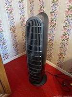 Tall black tower floor fan seen from front and side angles showing vertical grille, curved top controls, circular base, and power cord.
