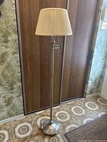 Image of the brass floor lamp standing on a patterned floor against a wood-paneled wall. The lamp has a pleated cream fabric shade, round metallic base, and slender brass pole.