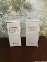 Two Miss Dior perfume boxes side by side on dark wooden surface with green and white floral background