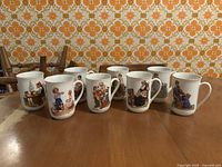 Eight white ceramic mugs with varied child and family scenes illustrated on each with colored Norman Rockwell style artwork and gold rims, sitting on wood table with orange floral wallpaper background.