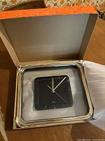 Quartz wall clock in original box showing black clock face, gold hands and markers, and square gold-tone frame.