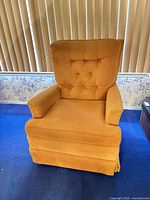 Mustard yellow retro armchair with button-tufted backrest, cushioned armrests, pleated skirt base, placed on blue carpet with vertical blinds in the background.