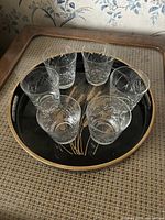 Six clear crystal etched glasses arranged in a circle on a black lacquered serving tray with a gold rim and decorative leaf/grass design.