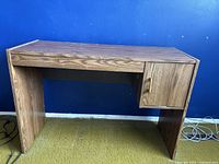 Photo showing a small rectangular wood finish desk with one storage drawer on the right side against a blue wall and green carpet.