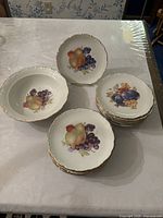 Set of 15 vintage fruit plates stacked and one fruit bowl, all decorated with colorful fruit motifs and gold trim edges.