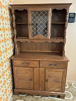 Photo of a medium brown wood hutch with upper shelves, central glass cabinet with lattice, two drawers and two doors below, standing against a wall with floral wallpaper and a tile floor.