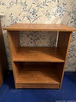 Single wood side table with two open shelves placed against floral wallpaper and blue carpet.